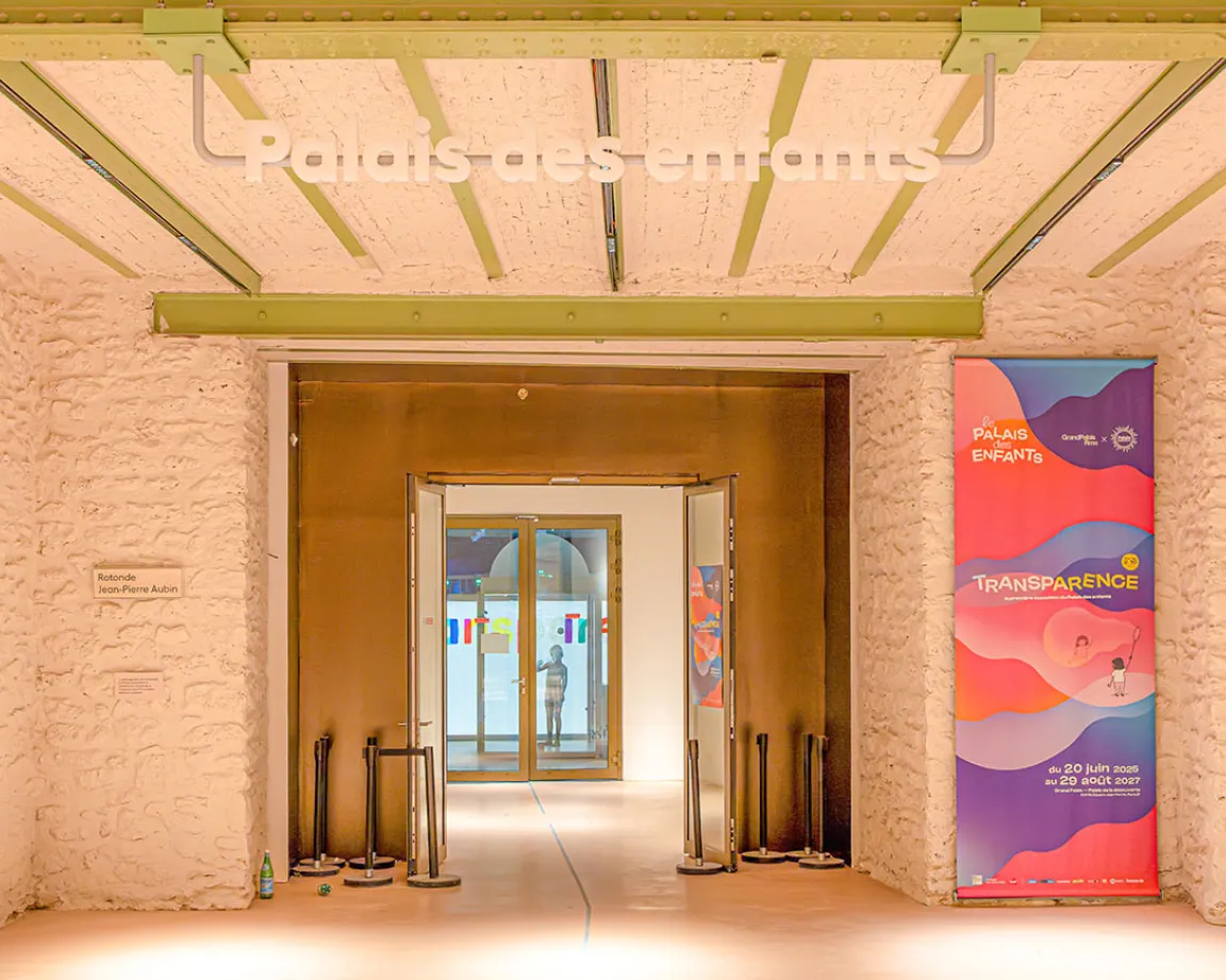 Vue de l'entrée du Palais des enfants avec une affiche de l'exposition "Transparence". On distingue au fond une silhouette, qui est la première sculpture visible pour les visiteurs de l'exposition.