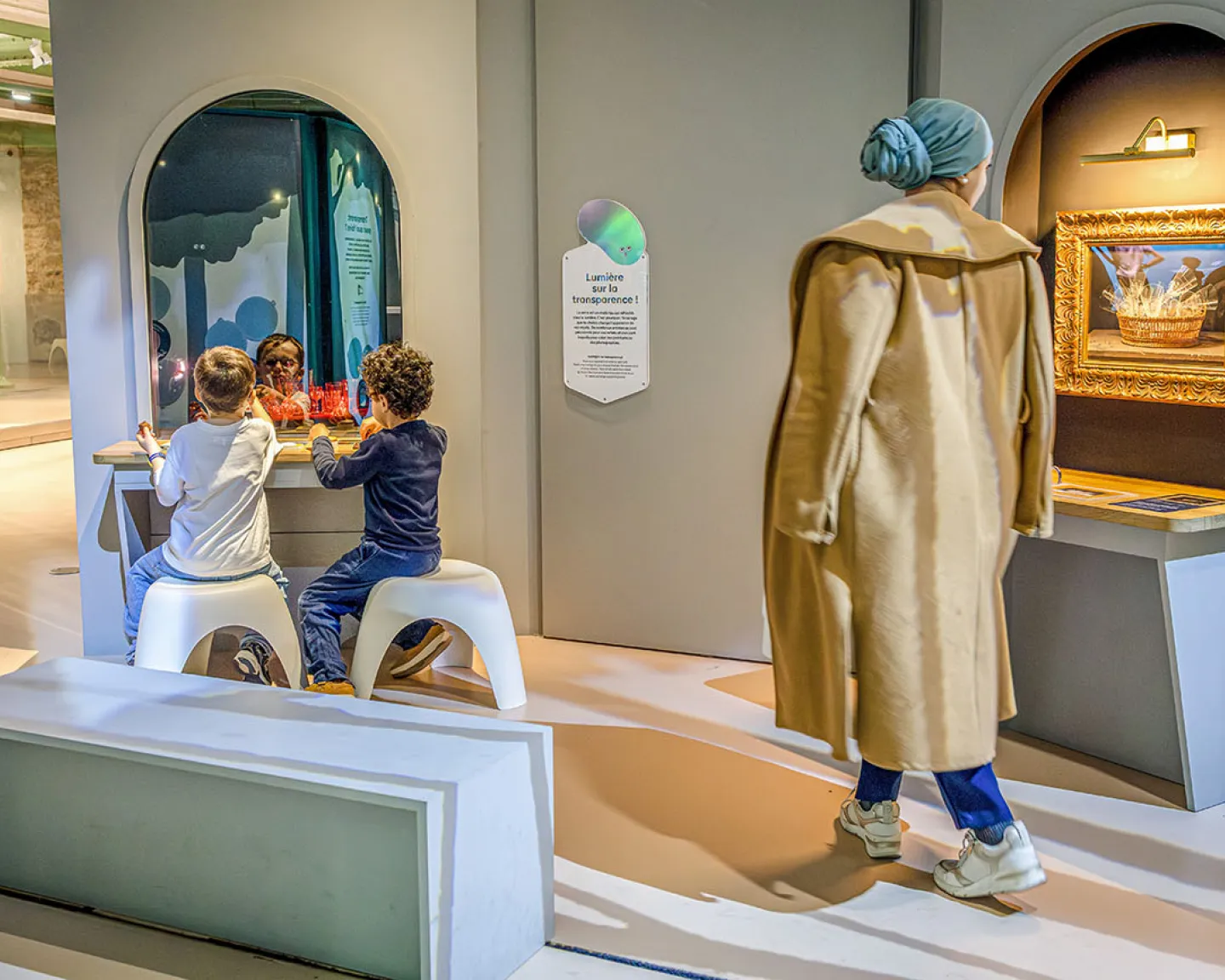 Vue intérieure du Palais des enfants avec une adulte débout et des enfants assis occupés à expérimenter un dispositif de l'exposition "Transparence"