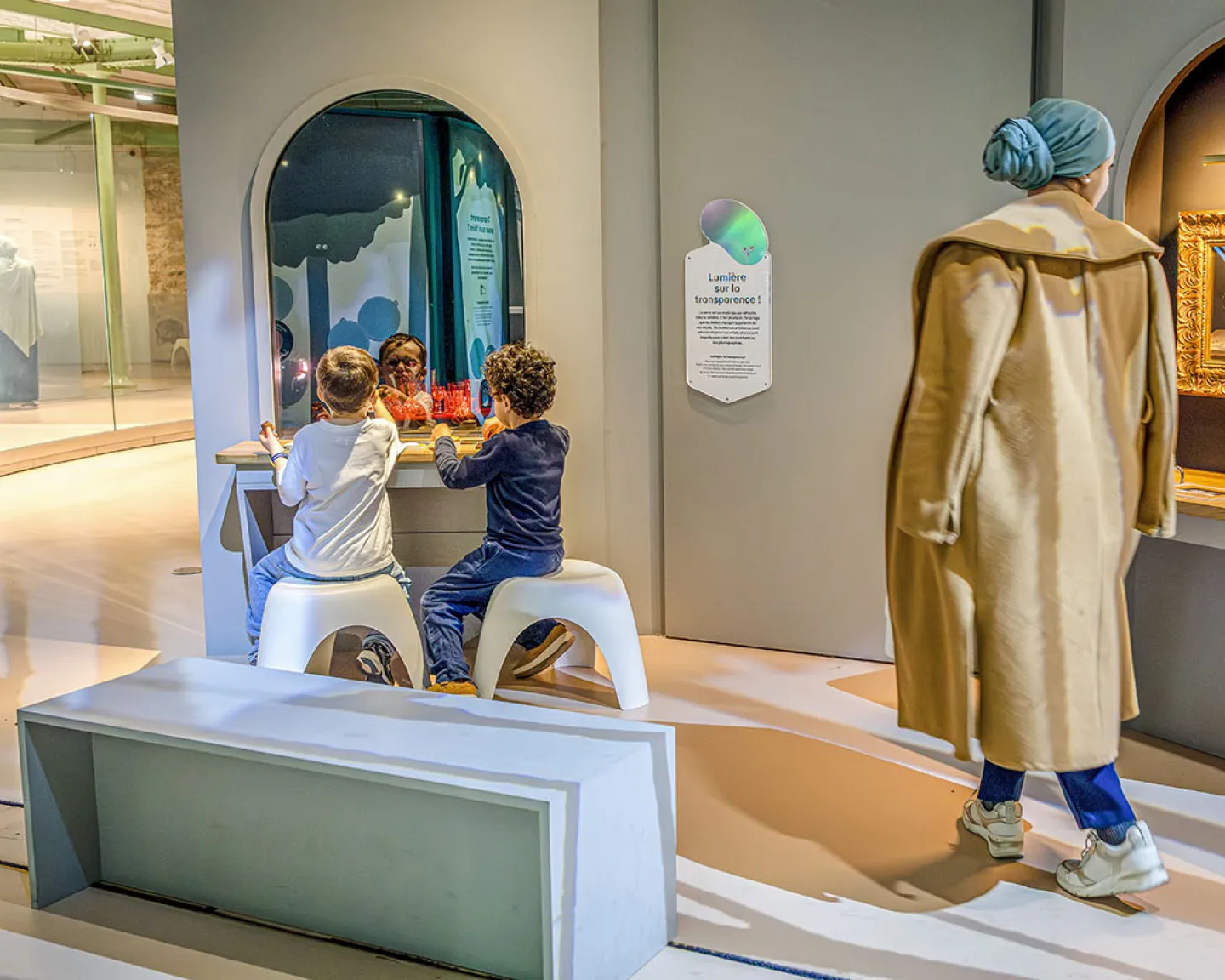 Vue intérieure du Palais des enfants avec une adulte débout et des enfants assis occupés à expérimenter un dispositif de l'exposition "Transparence"