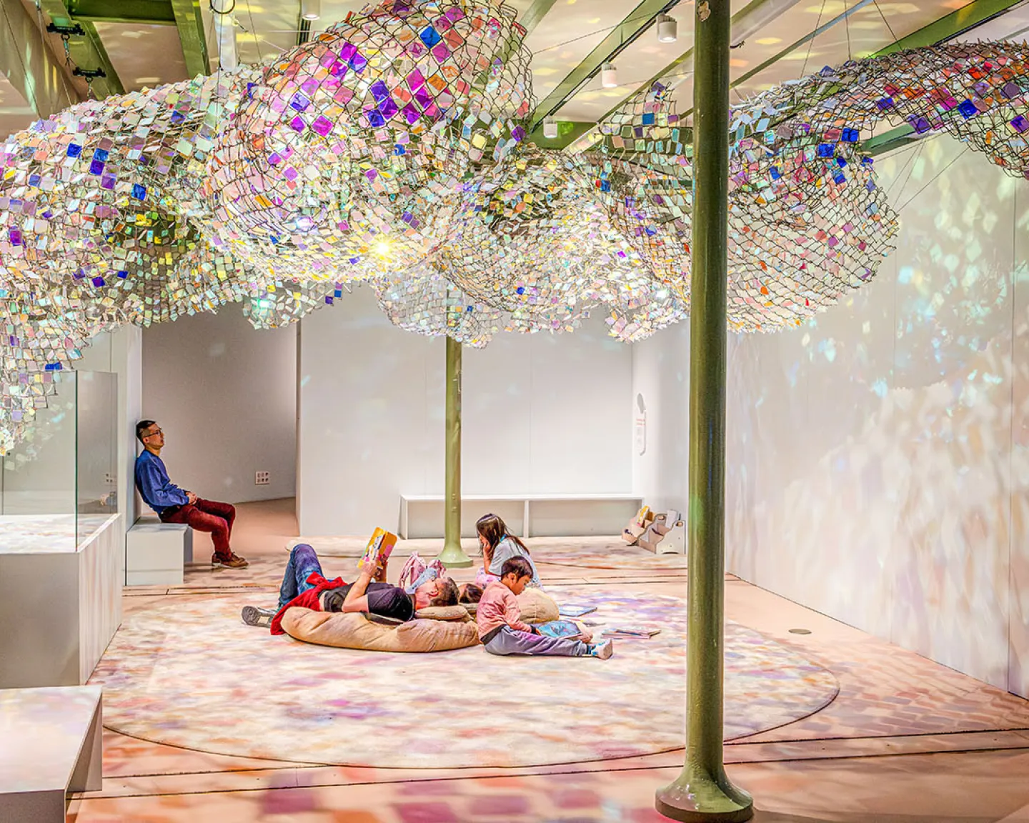 Vue intérieure du Palais des enfants, exposition Transparence, des enfants sont assis ou allongés et un adulte au loin est assis, sous l'œuvre Unwoven light de Soo Sunny Park